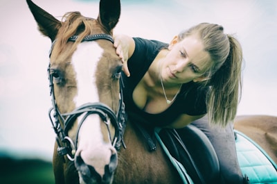 A woman with a ponytail gently leans forward to pet a brown horse with a white stripe on its face. The woman is wearing a black shirt and appears calm and attentive. The horse is equipped with a bridle, and there is a green saddle pad. The background is softly blurred, giving a serene and intimate atmosphere.