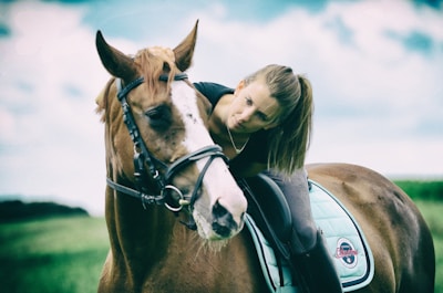 A person with long hair, wearing a black outfit, leans forward on top of a brown horse adorned with a bridle and a light blue saddle pad. The person appears to be engaging affectionately with the horse. The background showcases a blurred green field and a partly cloudy sky.