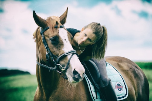 A person with long hair, wearing a black outfit, leans forward on top of a brown horse adorned with a bridle and a light blue saddle pad. The person appears to be engaging affectionately with the horse. The background showcases a blurred green field and a partly cloudy sky.