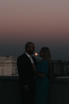 A silhouetted couple stands close together on a rooftop at dusk. The sky transitions from dark to a soft gradient of pink and grey. The moon is prominently visible in the background, casting a gentle glow over a distant urban landscape. The couple is partially obscured in shadow, creating a romantic and intimate atmosphere.
