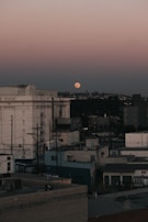 A cityscape at dusk featuring an industrial area with several large buildings and utility poles. The sky is a gradient from a light pink near the horizon to a deeper blue above. A large full moon is visible just above the horizon, adding a serene touch to the urban environment.
