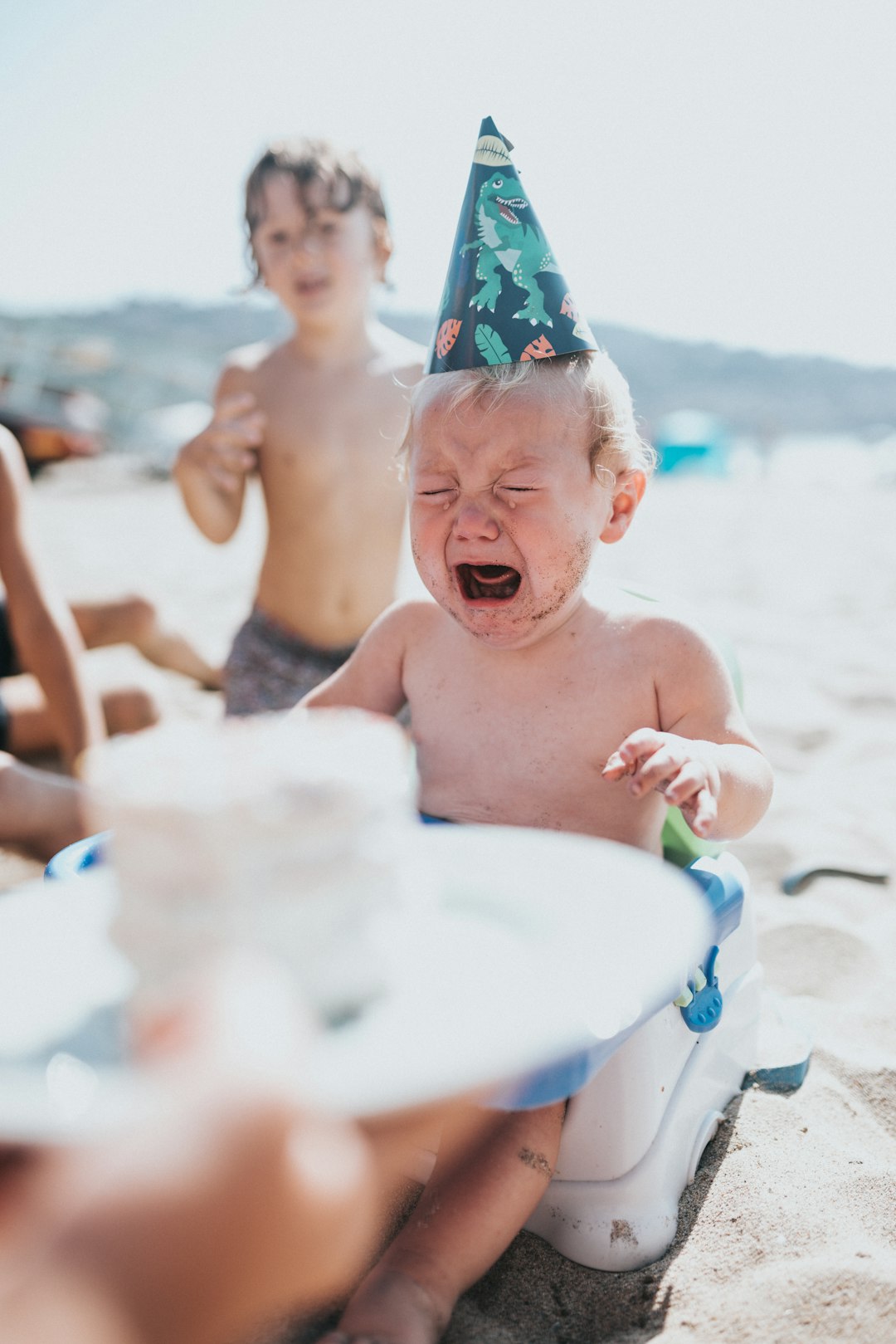 crying baby wearing dinosaur party hat during a birthday party on a beach. baby has either cake or sand or both smeared on face