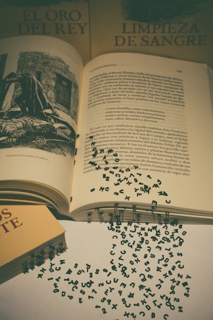 An open book with pages containing printed text and a black and white illustration is surrounded by two closed books that have titles in Spanish. Scattered around the books are small black letters, seemingly detached from the text, lying on a light-colored surface and creating an abstract and artistic arrangement.