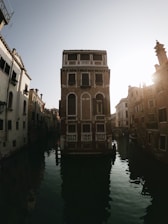 A tall, narrow building with a brick facade stands at the center of a canal, surrounded by other historic buildings. The early morning or late afternoon sun casts warm light, creating long shadows on the water. The architecture features classical Venetian elements with arched windows and decorative balconies.