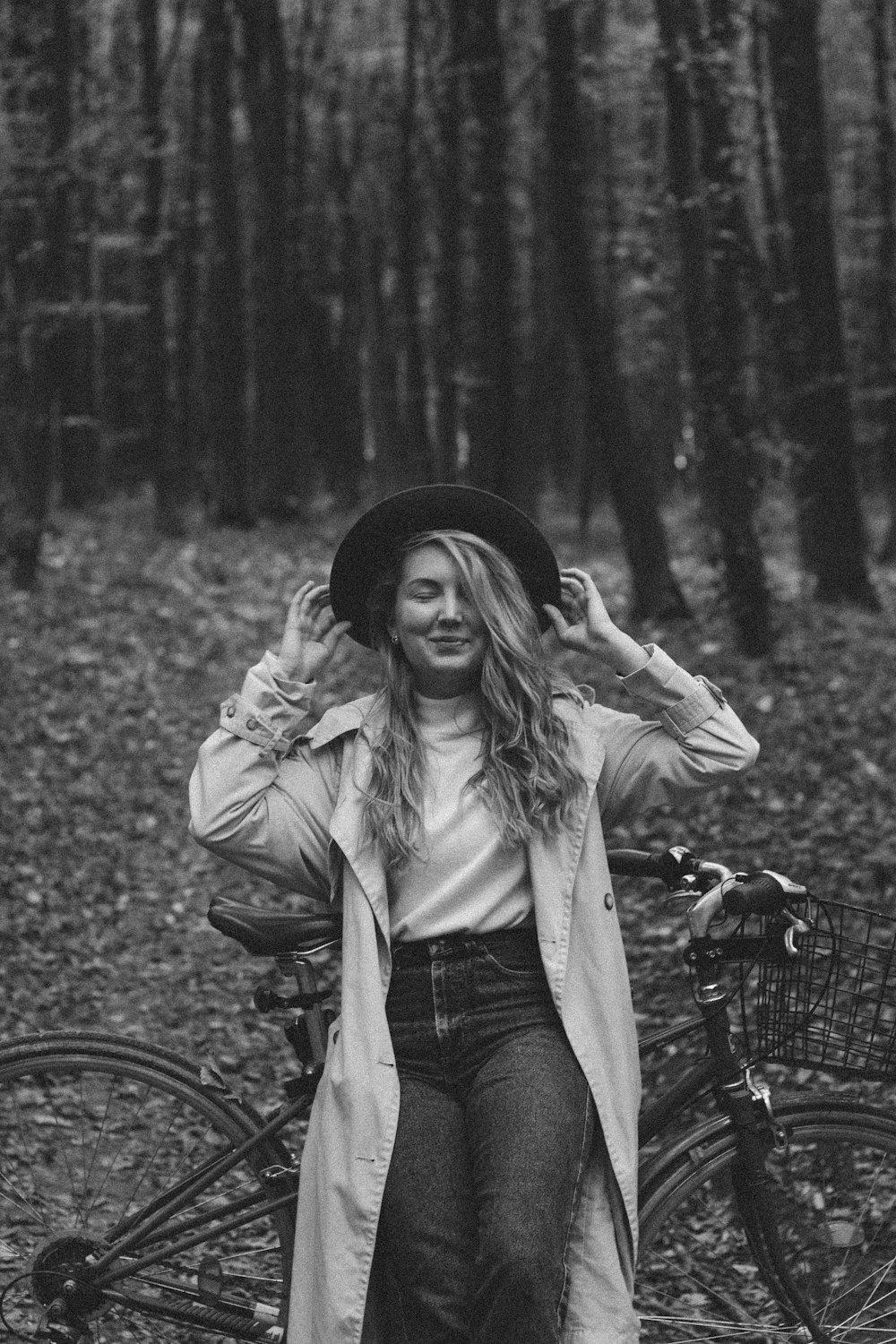 grayscale photo of woman in jacket and denim jeans standing beside bicycle