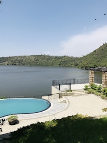 A serene lakeside view featuring a clear blue infinity pool overlooking a calm lake, surrounded by lush green hills. A stone patio with ornate railings and stone columns adds to the scenic beauty. Two birds can be seen flying in the clear blue sky above, and there is greenery and potted plants adorning the patio area.
