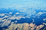 An aerial view of rugged mountain terrain with sharp ridges and valleys, partially covered by shadows and illuminated by sunlight. The terrain showcases a variety of shapes and patterns, with a gradient from light to dark as the landscape extends into the distance.