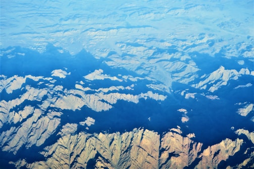 An aerial view of rugged mountain terrain with sharp ridges and valleys, partially covered by shadows and illuminated by sunlight. The terrain showcases a variety of shapes and patterns, with a gradient from light to dark as the landscape extends into the distance.