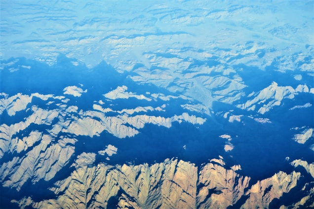 An aerial view of rugged mountain terrain with sharp ridges and valleys, partially covered by shadows and illuminated by sunlight. The terrain showcases a variety of shapes and patterns, with a gradient from light to dark as the landscape extends into the distance.
