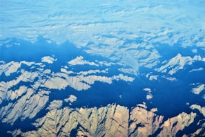 An aerial view of rugged mountain terrain with sharp ridges and valleys, partially covered by shadows and illuminated by sunlight. The terrain showcases a variety of shapes and patterns, with a gradient from light to dark as the landscape extends into the distance.