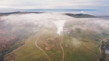 Aerial view of a lush, green landscape with rolling hills partially obscured by low-hanging clouds. Visible streams of steam rise from the ground, hinting at geothermal activity. The area features winding dirt roads and scattered patches of forest with autumnal colors.