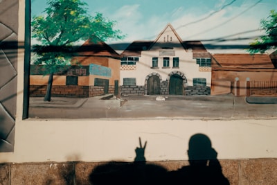 A mural of a building with arched doorways and windows, accompanied by a tree on the left side. There is a shadow of a person making a peace sign in the foreground.