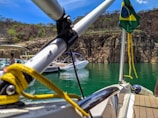 Boats are anchored on a green lake against a backdrop of rocky cliffs and sparse vegetation. A Brazilian flag is visible on one of the boats, and there are people relaxing on the watercraft.