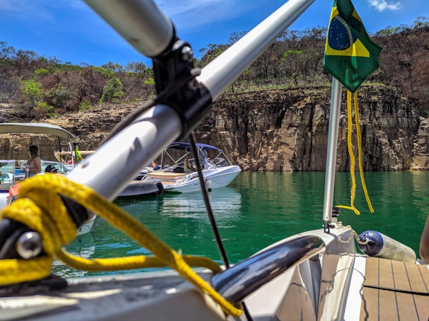 Boats are anchored on a green lake against a backdrop of rocky cliffs and sparse vegetation. A Brazilian flag is visible on one of the boats, and there are people relaxing on the watercraft.