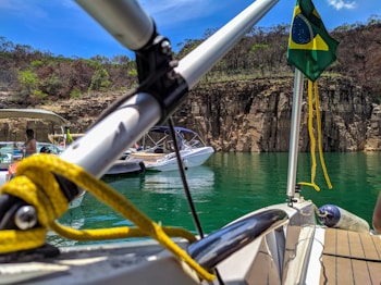Boats are anchored on a green lake against a backdrop of rocky cliffs and sparse vegetation. A Brazilian flag is visible on one of the boats, and there are people relaxing on the watercraft.
