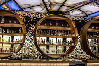 A stylish and modern bar display featuring circular wooden shelves filled with various bottles of alcohol. The background is an artistic, abstract pattern with reflective and metallic elements. Above, a decorative stained glass ceiling adds a touch of elegance and color.