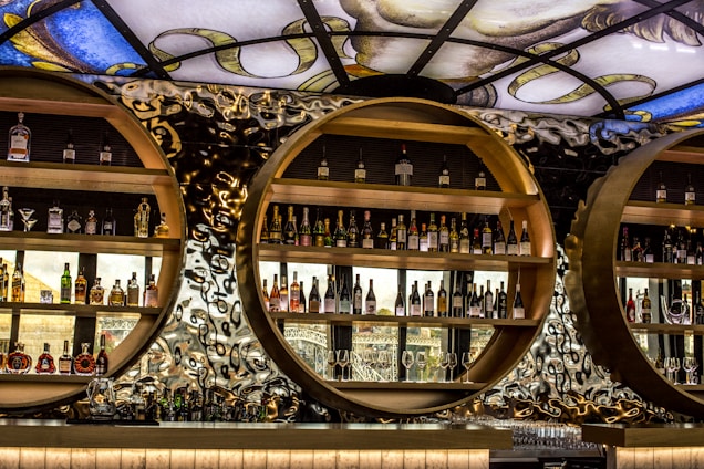 A stylish and modern bar display featuring circular wooden shelves filled with various bottles of alcohol. The background is an artistic, abstract pattern with reflective and metallic elements. Above, a decorative stained glass ceiling adds a touch of elegance and color.