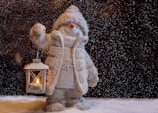 A snow-covered figure resembling a snowman dressed in a cozy coat and hat, holding a lantern with a lit candle. The scene is set amidst gently falling snowflakes, creating a cold yet warm ambiance with the glow of the lantern.