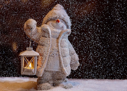 A snow-covered figure resembling a snowman dressed in a cozy coat and hat, holding a lantern with a lit candle. The scene is set amidst gently falling snowflakes, creating a cold yet warm ambiance with the glow of the lantern.