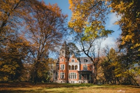 A picturesque, old brick mansion nestled among tall trees in a seasonal autumn setting. The leaves on the trees display various shades of yellow, orange, and brown, creating a warm and inviting atmosphere. A clear blue sky peeks through the branches, enhancing the vibrant scene.