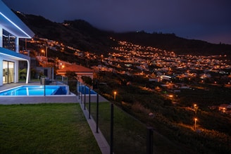 A modern house with illuminated windows and a pool sits on a hill overlooking a valley filled with numerous houses that are lit up under a dark evening sky. The scene is tranquil with a backdrop of mountains and vegetation.