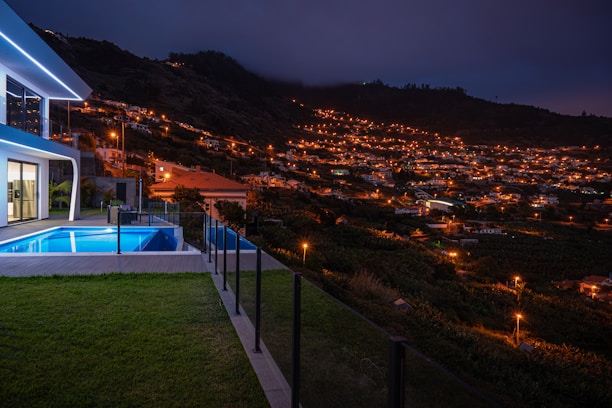 A modern house with illuminated windows and a pool sits on a hill overlooking a valley filled with numerous houses that are lit up under a dark evening sky. The scene is tranquil with a backdrop of mountains and vegetation.