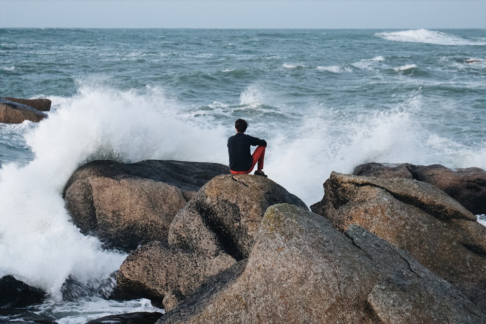 낮에는 바다 근처의 갈색 바위에 앉아 있는 검은색과 빨간색 긴팔 셔츠를 입은 남자