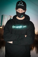 A person wearing a black cap, mask, and hoodie with 'StreamBloom' logos, stands with arms crossed and eyes closed. The background features a blurred cityscape and a body of water, with a teal hue dominating the scene.