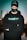 A person wearing a black cap, mask, and hoodie with 'StreamBloom' logos, stands with arms crossed and eyes closed. The background features a blurred cityscape and a body of water, with a teal hue dominating the scene.