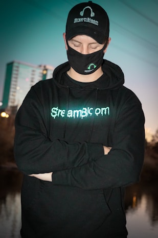 A person wearing a black cap, mask, and hoodie with 'StreamBloom' logos, stands with arms crossed and eyes closed. The background features a blurred cityscape and a body of water, with a teal hue dominating the scene.