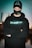 A person wearing a black cap, mask, and hoodie with 'StreamBloom' logos, stands with arms crossed and eyes closed. The background features a blurred cityscape and a body of water, with a teal hue dominating the scene.
