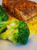 A piece of seasoned salmon sits atop a bed of golden saffron rice with a side of steamed broccoli. The salmon is richly coated with spices, giving it a textured and flavorful appearance. The broccoli appears vibrant and freshly cooked, adding a touch of green to the dish.