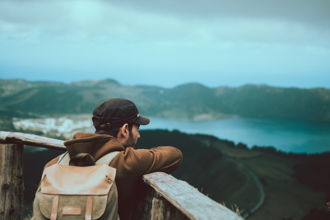 man in brown jacket and black cap sitting on concrete wall looking at the sea during