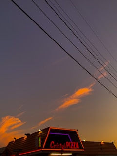 A small pizza restaurant with illuminated signage during sunset. The sky is a mix of dark blue and orange hues with a few clouds. Several power lines stretch across the sky, adding an urban feel.