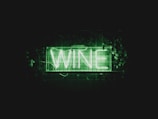 A neon sign displays the word 'WINE' in bright green letters. The sign is placed against a dark background and is surrounded by green foliage and leaves.