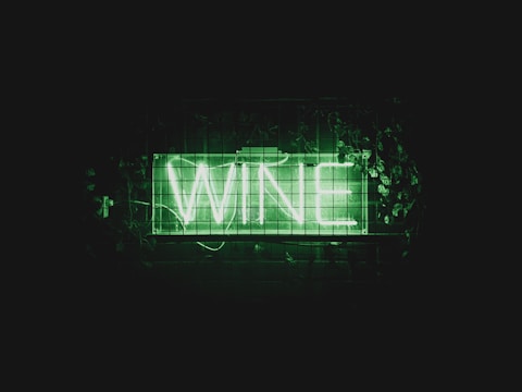 A neon sign displays the word 'WINE' in bright green letters. The sign is placed against a dark background and is surrounded by green foliage and leaves.