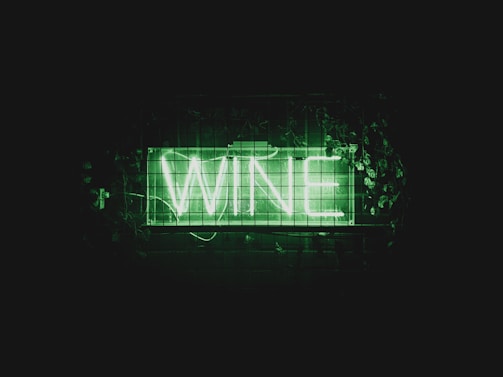 A neon sign displays the word 'WINE' in bright green letters. The sign is placed against a dark background and is surrounded by green foliage and leaves.