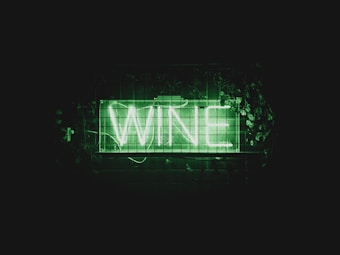 A neon sign displays the word 'WINE' in bright green letters. The sign is placed against a dark background and is surrounded by green foliage and leaves.