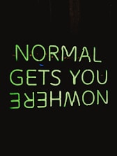 A neon sign with the phrase 'NORMAL GETS YOU NOWHERE' written in bright green against a black background. The neon text is reflected upside down under the original phrase.