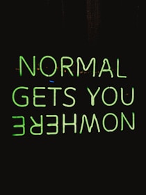 A neon sign with the phrase 'NORMAL GETS YOU NOWHERE' written in bright green against a black background. The neon text is reflected upside down under the original phrase.