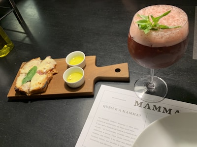 A wooden serving board holds slices of bread topped with melted cheese and a basil leaf. Beside the bread, there are two small white ramekins with yellow sauce. A frothy pink cocktail garnished with green leaves is in a large glass nearby. Underneath, there is a menu partially visible on the table, labeled with 'MAMMA.'