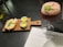 A wooden serving board holds slices of bread topped with melted cheese and a basil leaf. Beside the bread, there are two small white ramekins with yellow sauce. A frothy pink cocktail garnished with green leaves is in a large glass nearby. Underneath, there is a menu partially visible on the table, labeled with 'MAMMA.'