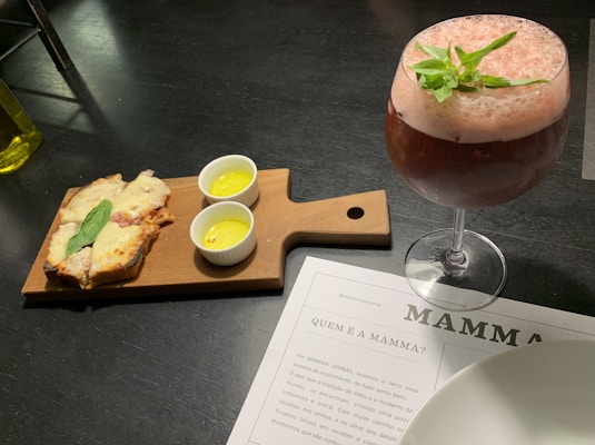 A wooden serving board holds slices of bread topped with melted cheese and a basil leaf. Beside the bread, there are two small white ramekins with yellow sauce. A frothy pink cocktail garnished with green leaves is in a large glass nearby. Underneath, there is a menu partially visible on the table, labeled with 'MAMMA.'