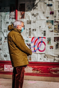 An elderly person wearing a face mask and a green coat stands in front of a wall covered with newspaper pages and various images. The individual appears to be observing something off-camera and the scene is brightly lit by sunlight.
