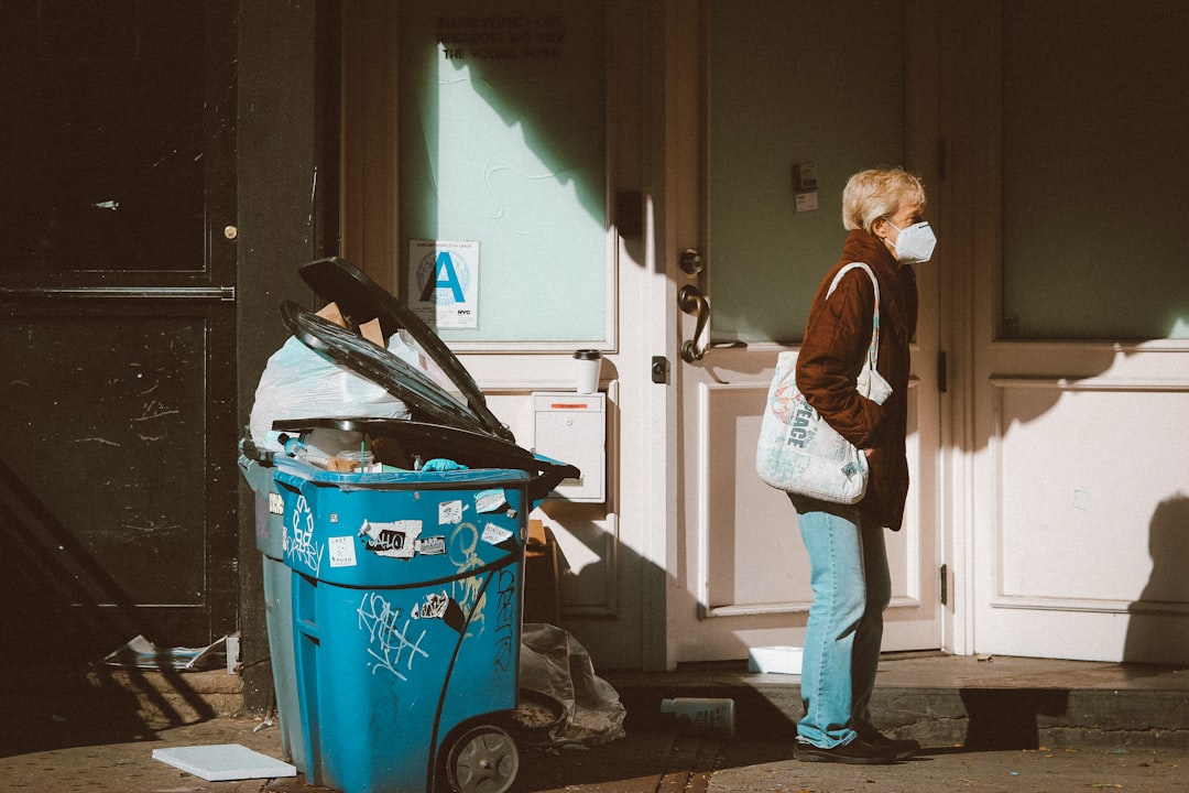 woman in brown jacket and blue denim jeans standing beside red plastic trash bin