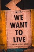 A poster with bold black text reading 'WE WANT TO LIVE' against a backdrop of environmental and social issues listed around the edges. The top features the logo and name 'extinction rebellion.' The design uses contrasting colors and typography to emphasize urgency and activism.