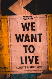 A poster with bold black text reading 'WE WANT TO LIVE' against a backdrop of environmental and social issues listed around the edges. The top features the logo and name 'extinction rebellion.' The design uses contrasting colors and typography to emphasize urgency and activism.