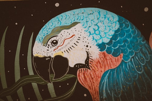 A vibrantly colored painting of a parrot with predominantly blue, white, and red feathers against a dark backdrop with a few scattered small white circles. The parrot is holding a green leaf in its beak.