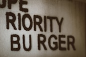 Text appears handwritten on a textured wall, featuring the words 'PRIORITY BURGER' in uppercase letters. The text is bold and black against a light gray or off-white background.