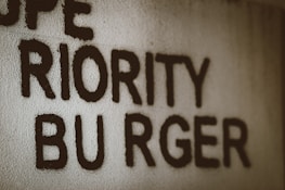 Text appears handwritten on a textured wall, featuring the words 'PRIORITY BURGER' in uppercase letters. The text is bold and black against a light gray or off-white background.